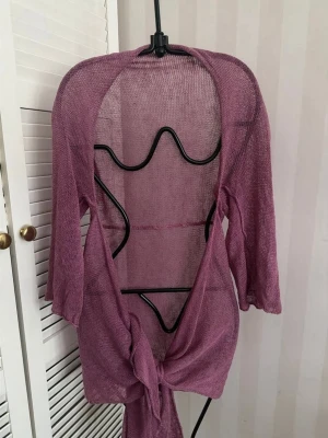 Ljuslila stickad cardigan - Säljer en tunnstickad, ljuslila cardigan med trekvartsärmar. Modellen är öppen framtill och kan knytas i midjan för en avslappnad look. Materialet är lätt och genomskinligt, perfekt för lager på lager-stil.