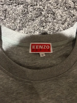 Grå Kenzo sweatshirt med tiger - Säljer en grå sweatshirt från Kenzo med ikoniskt tigertryck och broderad logga på bröstet. Klassisk rund halsringning och ribbade muddar. Perfekt för dig som vill ha en statementtröja med streetvibe.