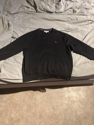 Svart sweatshirt från Polo  - Klassisk svart sweatshirt från Polo med rund halsringning och diskret röd logga broderad på bröstet. Tröjan har ribbade muddar vid ärmslut och nederkant. Perfekt för en avslappnad och stilren look.
