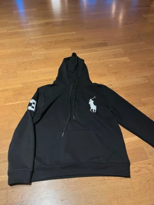 Svart hoodie från Polo Ralph Lauren - Svart hoodie från Polo Ralph Lauren med stor vit polospelare broderad på bröstet och en vit siffra 3 på ena ärmen. Klassisk känguruficka framtill och justerbar huva med snören. Tillverkad i mjuk bomullsblandning för skön känsla. Storlek M men passar Xs-Xxs. Mått: 58cm längd, 45cm bredd. För nån runt 155-165cm.