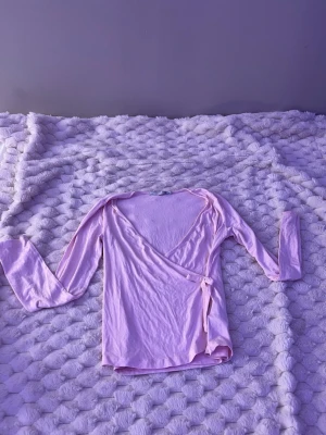 Ljusrosa omlottopp med knyt - Supersöt ljusrosa omlottopp med knyt i sidan och v-ringad hals. Toppen har långa ärmar och är gjord i ett mjukt, tunt material som känns skönt mot huden. Perfekt att styla med jeans eller kjol för en trendig look.