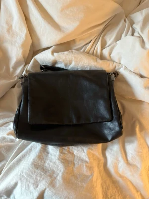 Svart axelväska i läder med kedja - Snygg svart axelväska i mjukt läder med bred lockstängning och silverfärgade kedjedetaljer på axelremmen. Väskan har en stilren och enkel design som passar till det mesta. Perfekt för dig som gillar en klassisk men trendig look. Från Zara.