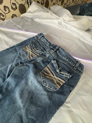 Blå bootcut jeans med broderade fickor mudd jeans - Säljer ett par blå bootcut jeans med snygga broderade detaljer på bakfickorna i flera färger. Jeansen har låg midja, slitningar framtill och klassisk femficksmodell. Perfekta för dig som gillar en retro och avslappnad stil.