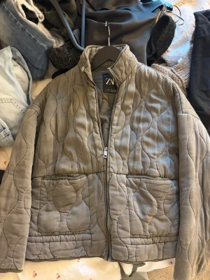 Grå quiltad cardiganjacka från ZARA - Quiltad grå cardiganjacka från ZARA med vågigt mönster, dragkedja framtill och stora fickor. Jackan har en hög krage. Välanvänd men bra skick, bilder visar lätt slitage