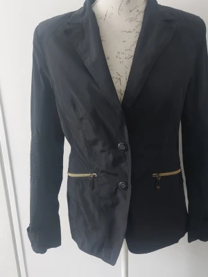 Svart kavaj med dragkedjedetaljer - Stilren svart kavaj med två knappar framtill och guldfärgade dragkedjor vid fickorna. Figurnära passform och detaljerade sömmar på ärmarna. Materialet är tunt och lätt, perfekt för en trendig look. Passar till många olika outfits.