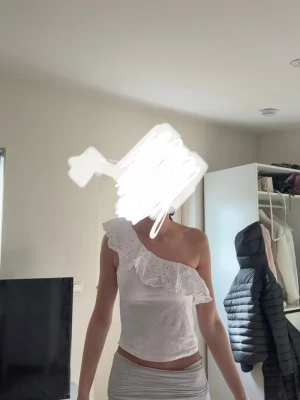 Vit oneshoulder topp med volang - Säljer en vit oneshoulder topp med bred volangkant och broderade detaljer. Toppen är figurnära och har en romantisk känsla med sin asymmetriska axel och mjuka material. Perfekt för dig som vill sticka ut lite extra.