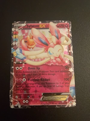 Sylveon EX Pokémonkort - Säljer ett Sylveon EX samlarkort från Pokémon TCG. Kortet har en glittrig rosa design med motiv av Sylveon och Eevee. Det är ett EX-kort med 170 HP och attacker som Dress Up och Precious Ribbon. Perfekt för samlare eller Pokémonfans!