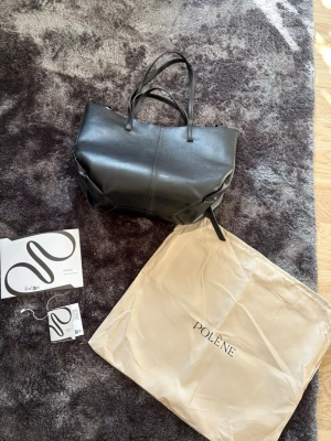Svart handväska från Polène - Snygg svart handväska från Polène i slätt läder med två handtag små handtag och 2 lite längre och minimalistisk design. Väskan har diskret Polène-logga i guld framtill och kommer med original dustbag och tags. Perfekt för dig som gillar stilrena accessoarer. I ett av spännena har en söm lossnat, man kan antigen sy eller limma men påverkar egentligen inte funktionen utan det går bra att stänga den ändå! 💕🌸