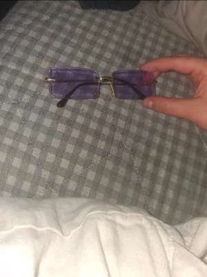 Lila rektangulära solglasögon - Säljer ett par trendiga solglasögon med rektangulära, lila glas och tunna guldfärgade bågar. Glasögonen har en minimalistisk design och svarta skalmar. Perfekta för dig som vill sticka ut med en cool accessoar.