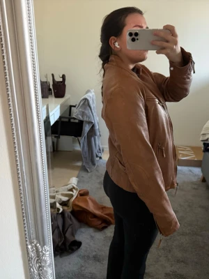 Brun skinnjacka med dragkedjor - Snygg brun skinnjacka med ståkrage och dragkedjor framtill samt på fickorna. Jackan har en klassisk bikerstil och sitter figurnära. Perfekt för dig som vill ha en tidlös och cool look.