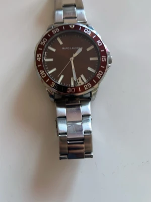 Marc Lauder silverfärgad klocka - Stilren klocka från Marc Lauder med silverfärgad länk i metall och mörkröd urtavla med vita detaljer. Klockan har tydliga siffror runt boetten och datumvisning. Perfekt accessoar för dig som gillar en klassisk men modern look. (OBSERVANT klockan är sönder inuti men det går att fixa)