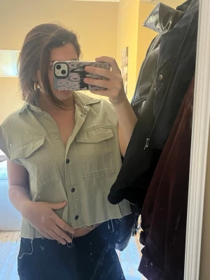 Grön croppad skjorttopp med knappar - Säljer en grön croppad skjorttopp med knappar framtill och två bröstfickor. Toppen har korta ärmar och rå, fransig nederkant som ger en cool look 