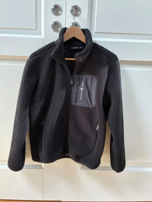 Svart teddyjacka från Sail Racing - Mysig svart teddyjacka från sail racing med hög krage och dragkedja framtill. Jackan har en bröstficka med logga och två sidofickor med dragkedja. Perfekt för kyliga dagar till höst och vår och enkel att matcha med olika outfits.