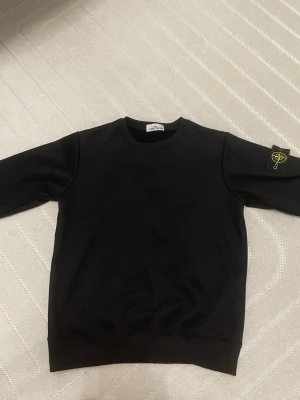 Svart sweatshirt från Stone Island - Säljer en svart sweatshirt från Stone Island med klassisk rund hals och ribbade muddar. Tröjan har den ikoniska Stone Island-loggan på vänster ärm. Perfekt för dig som gillar streetwear och enkel stil.