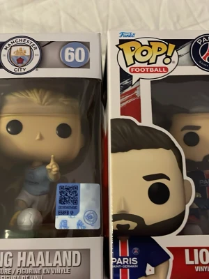 Funko Pop! fotbollsfigurer Haaland & Messi - Två Funko Pop! vinylfigurer: Erling Haaland i Manchester Citys ljusblå matchställ med boll och Lionel Messi i Paris Saint-Germains mörkblå dräkt. Båda figurerna är i sina originalkartonger och har tydliga klubbmärken på förpackningarna.