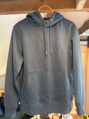 Blå hoodie från Tommy Hilfiger - Snygg blå hoodie från Tommy Hilfiger med klassisk logga på bröstet och randig detalj på huvan. Hoodien har känguruficka framtill, dragsko i huvan och ribbade muddar. Perfekt för en chill och stilren look.