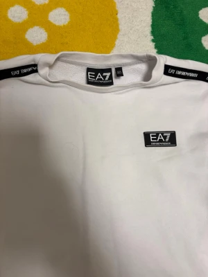 Vit EA7 sweatshirt med logga - Snygg vit sweatshirt från EA7 Emporio Armani med svart logga på bröstet och svarta band med EA7-tryck längs ärmarna. Tröjan har rund halsringning och är tillverkad i mjukt material som känns skönt mot huden. Perfekt för en clean och sportig look.