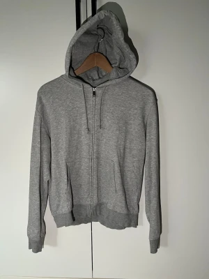 Grå zip hoodie med huva - Klassisk grå hoodie med dragkedja framtill och två fickor. Tröjan har justerbar huva med snören och ribbade muddar vid ärmar och nederkant. Perfekt för en avslappnad och stilren look. Tillverkad i mjukt sweatshirtmaterial. Storlek xxs, men skulle säga att den passar xs