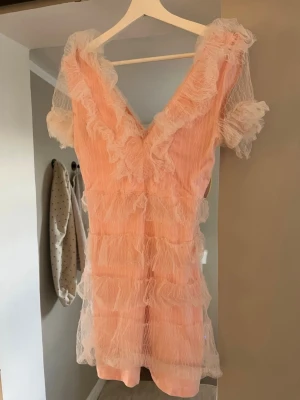 Ljusrosa volangklänning med tyll - Supersöt ljusrosa kortklänning med volanger och transparent tyll. Djup v-ringning både fram och bak samt korta puffärmar. Klänningen har flera lager tyll och ger en romantisk och lekfull vibe. Perfekt för dig som vill sticka ut med en unik look!