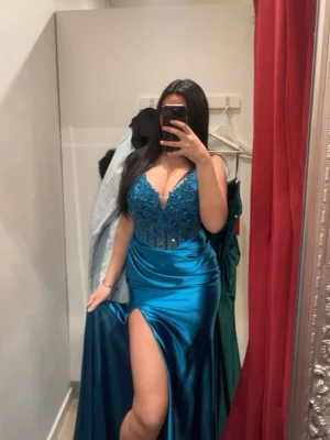 Blå balklänning med slits och spets - Säljer en glansig blå långklänning med djup v-ringning, spetsdetaljer och pärlor upptill. Klänningen har smala axelband, markerad midja och en hög slits framtill. Perfekt för bal eller festkvällar.