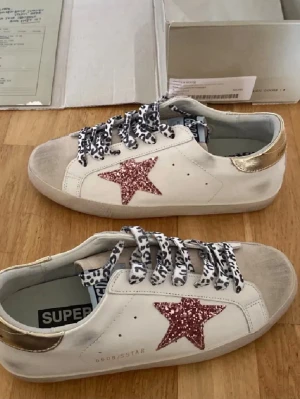 Golden Goose sneakers med glitterstjärna - Snygga vita Golden Goose sneakers med rosa glitterstjärna på sidan, guldfärgad häl och svartvita leopardmönstrade skosnören. Jag säljer dom för billigt då jag vill bli av med dom men också att jag bytt stil!