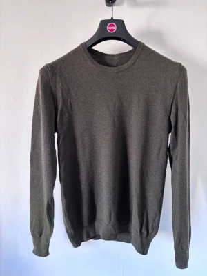 J.lindeberg stickad tröja i merinoull  - En stilren olivgrön stickad tröja med rund halsringning och långa ärmar. Tröjan har ribbade muddar vid ärmslut och nederkant. Perfekt att matcha med jeans eller chinos för en enkel och snygg look.