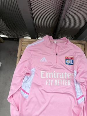  Olympique Lyonnais träningströja - Snygg rosa träningströja från Adidas med Olympique Lyonnais klubbmärke och Emirates Fly Better-tryck på bröstet. Tröjan har halv dragkedja, vita detaljer på sidorna och axlarna samt lång ärm. Perfekt för fotbollsträning eller chill.