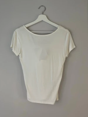 Vit topp med satinrosett - Snygg vit topp med korta ärmar och rund halsringning. Baksidan har en djupare urringning med en dekorativ satinrosett som detalj. Tillverkad i mjukt och lätt material, perfekt för en clean och stilren look.