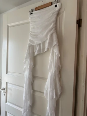 Toppi S - Säljer en vit kjol med asymmetrisk design och långa volanger i flera lager. Kjolen är gjord av ett tunt, skirt tyg som ger ett flowy och unikt intryck. Perfekt för dig som vill ha en kreativ och trendig stil.