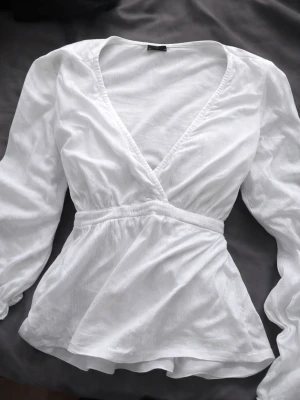Vit omlottblus med knytband - Stilren vit omlottblus med långa ärmar och knytband i midjan. Blusen har en djup v-ringning och är tillverkad i ett lätt, något strukturerat tyg som ger en luftig känsla. Perfekt att styla med jeans eller kjol för en sommrig look.