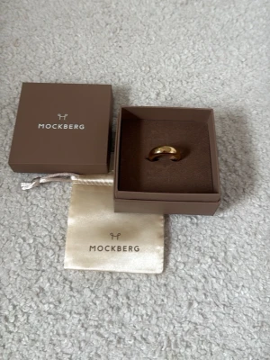 Guldfärgad ring från Mockberg storlek 56 - Snygg guldfärgad ring från Mockberg i stilren och klassisk design. Ringen har en slät, rundad form och levereras i original ask och tygpåse med Mockberg-logga. Perfekt accessoar för dig som gillar minimalistisk och elegant stil. Nypris 599