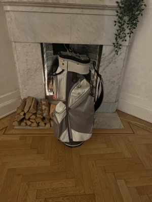 Grå golfbag med flera fack - Snygg golfbag i grått och vitt med flera praktiska fack och avdelare för klubbor. Väskan har bärrem och många fickor för tillbehör. Perfekt för dig som vill ha ordning på golfbanan.