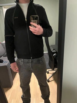 Svart zip hoodie från BOSS - Snygg svart hoodie från BOSS med dragkedja framtill och diskreta mönstrade detaljer längs axlar och ärmar. Klassisk passform med ribbade muddar och liten BOSS-logga på bröstet. Perfekt för en clean och stilren look.