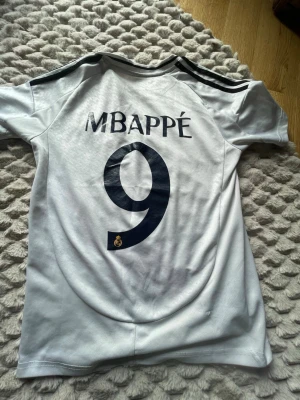 Vit Real Madrid Mbappé tröja - Snygg vit fotbollströja med Mbappé och nummer 9 på ryggen, samt Real Madrid-logga. Tröjan har korta ärmar och svarta detaljer på axlarna. Perfekt för dig som älskar fotboll och vill bära din favoritspelare på ryggen.