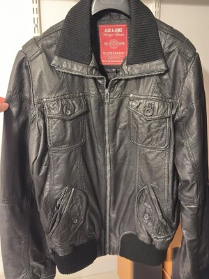 Svart skinnjacka Jack & Jones - Snygg svart skinnjacka från Jack & Jones med ribbad krage, muddar och nederkant. Jackan har fyra framfickor med lock och knapp, samt dragkedja framtill. Perfekt för dig som vill ha en klassisk men cool look.