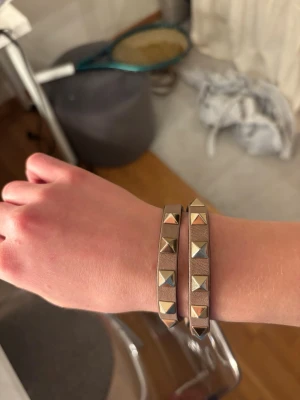 Valentino armband dubbelt - Snyggt brunt valentino armband i läder med dubbla rader av silvriga pyramidformade nitar. Armbandet har en modern och edgy look som passar perfekt till streetwear eller för att ge outfiten lite extra attityd. Justerbar längd för bästa passform från Valentino.  Pris kan diskuteras vid snabb affär