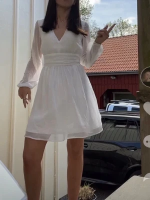Vit kort omlottklänning med v-ringning - Säljer en vit kort klänning med v-ringning och omlottdetalj. Klänningen har långa, transparenta ärmar och markerad midja som ger en snygg siluett. Perfekt för sommarens alla tillfällen! 💫