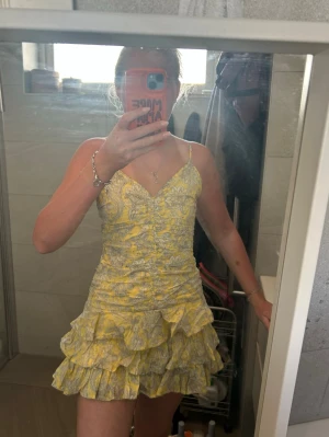 Gul blommig volangklänning - Söt kort gul klänning med blommigt mönster och volanger. Klänningen har smala axelband, v-ringning och omlottdetalj framtill. Perfekt för sommarens alla tillfällen och ger en fräsch och ungdomlig look.