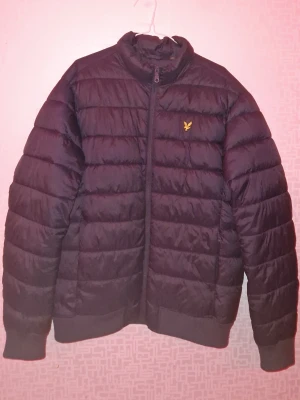 Svart pufferjacka från Lyle & Scott - Snygg svart pufferjacka från Lyle & Scott med gul logga på bröstet. Jackan har ståkrage, dragkedja framtill och ribbade muddar vid ärmslut och nederkant. Tillverkad i ett glansigt, quiltat material som håller värmen under kalla dagar.