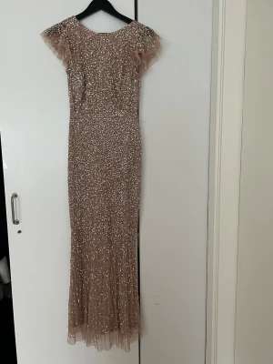 Glittrig champagnefärgad långklänning - Säljer en elegant långklänning i champagnefärg med glittriga paljetter över hela klänningen. Klänningen har korta volangärmar, rund halsringning fram och djup v-ringning i ryggen. Materialet är troligtvis polyester och klänningen har en figurnära passform.