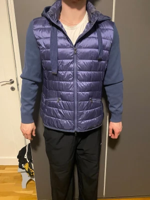 Blå Cardigan från Massimo Dutti - Snygg blå cardigan från Massimo Dutti med quiltad front och stickade ärmar. Jackan har huva med breda snören, två dragkedjeförsedda fickor och hel dragkedja framtill. Perfekt för dig som vill ha en modern och bekväm jacka med coola detaljer.