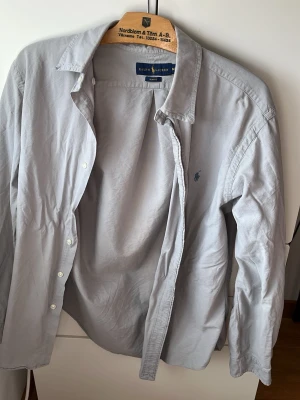 Grå slim fit skjorta från Ralph Lauren - Säljer en stilren grå skjorta från Ralph Lauren i slim fit-modell. Skjortan har klassisk krage, knappar framtill och den ikoniska loggan broderad på bröstet. Perfekt för dig som gillar en clean look och vill ha något som funkar till många outfits. Perfekt skick, och det är som att den aldrig blivit använd förut. Gäller bara att stryka den!