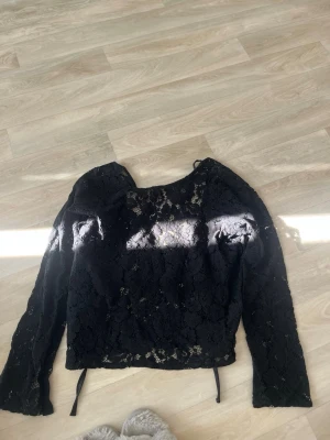 Svart spetsblus med knytband - Snygg svart blus i spets med lång ärm och rund halsringning. Blusen har transparent spetsmönster och knytband i ryggen för extra detalj. Perfekt att styla med höga jeans eller kjol för en trendig look.