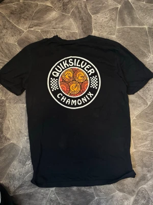 Svart Quiksilver t-shirt Chamonix - Svart t-shirt från Quiksilver med stort färgglatt tryck på ryggen där det står 'Quiksilver Chamonix' och har grafiska detaljer i rött, gult och vitt. Klassisk passform och rund hals. Perfekt för dig som gillar streetwear och snygga prints.