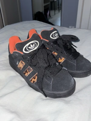 Adidas svarta sneakers med leoparddetaljer - Svarta Adidas sneakers med feta svarta skosnören, orange insida och leopardmönstrade stripes på sidorna. Skorna har en låg profil och är gjorda i mockaliknande material. Adidas-logga på plösen och cool streetstyle-vibe. Dom är aldrig använda och är helt nya.