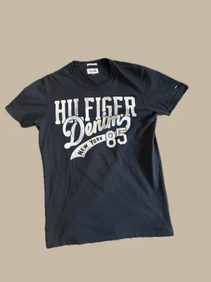 Svart Hilfiger Denim t-shirt - Snygg svart t-shirt från Hilfiger Denim med stort vitt tryck på bröstet där det står 'Hilfiger Denim New York 85'. Klassisk rund halsringning och korta ärmar. Perfekt till jeans för en avslappnad look.