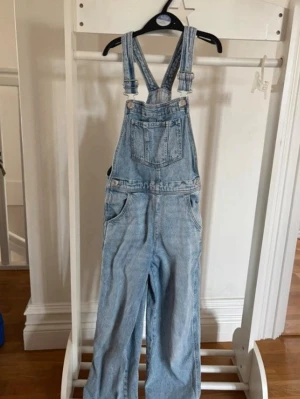 Ljusblå denim byxdress med hängslen - Säljer en ljusblå denim byxdress med klassiska justerbara hängslen och raka ben. Byxdressen har en stor bröstficka framtill samt sidofickor och bakfickor. Perfekt för en avslappnad och trendig look.