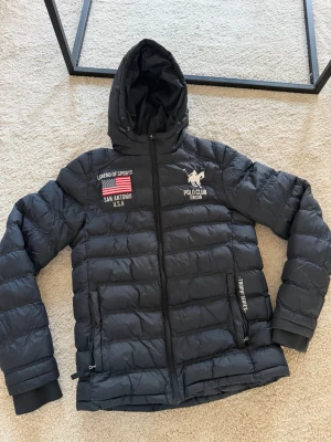 Svart pufferjacka Polo Club Vinson - Säljer en svart pufferjacka från Polo Club Vinson med broderad amerikansk flagga och texten 'Legend of Sports' samt 'San Antonio U.S.A' på bröstet. Jackan har huva, dragkedja och två sidofickor med dragkedja. Perfekt för kalla dagar