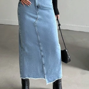 Lång jeanskjol - Säljer en snygg lång jeanskjol i ljusblå denim med rå, fransad kant nedtill och asymmetrisk söm framtill. Kjolen har klassisk femficksdesign och stängs med knapp och dragkedja. Perfekt att styla med boots eller sneakers.