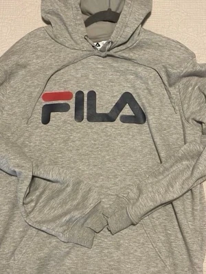 Grå hoodie från FILA med logga - Snygg grå hoodie från FILA med stor logga i rött och marinblått på bröstet. Klassisk modell med huva och ribbade muddar. Perfekt för en avslappnad streetwear-look. Tillverkad i mjukt och bekvämt material.